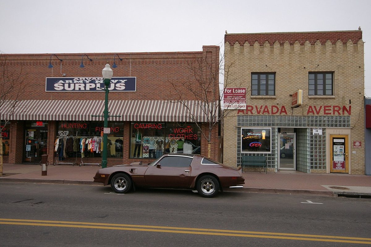 Brick storefronts in Olde Town Arvada, an army-navy surplus store and the Arvada Tavern, with a brown 1970s coupe parked at the curb