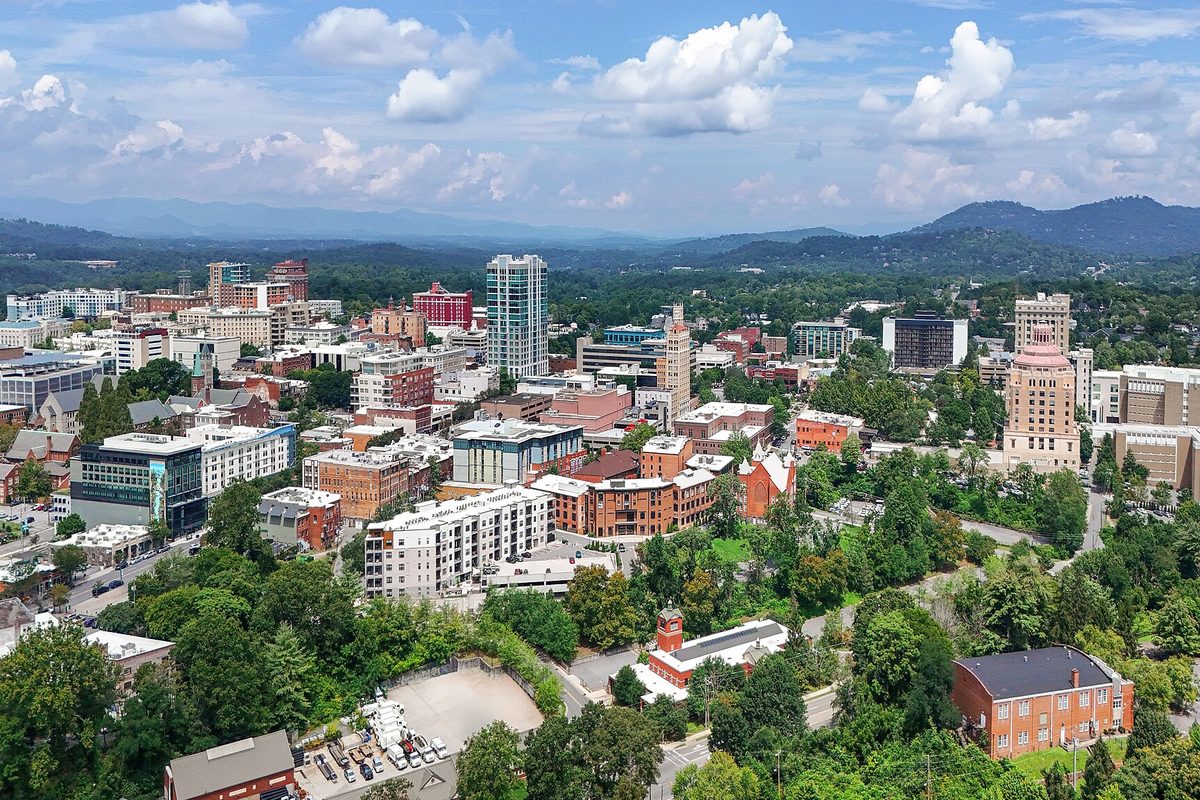 Aerial view of downtown Asheville, brick buildings and a few towers among green trees, with the Blue Ridge Mountains behind under a blue sky