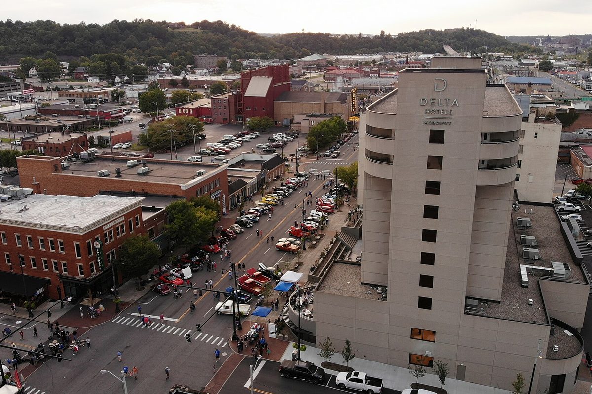Aerial view of downtown Ashland with a car show lining the street beside the round Delta Hotels tower, brick buildings and wooded hills behind