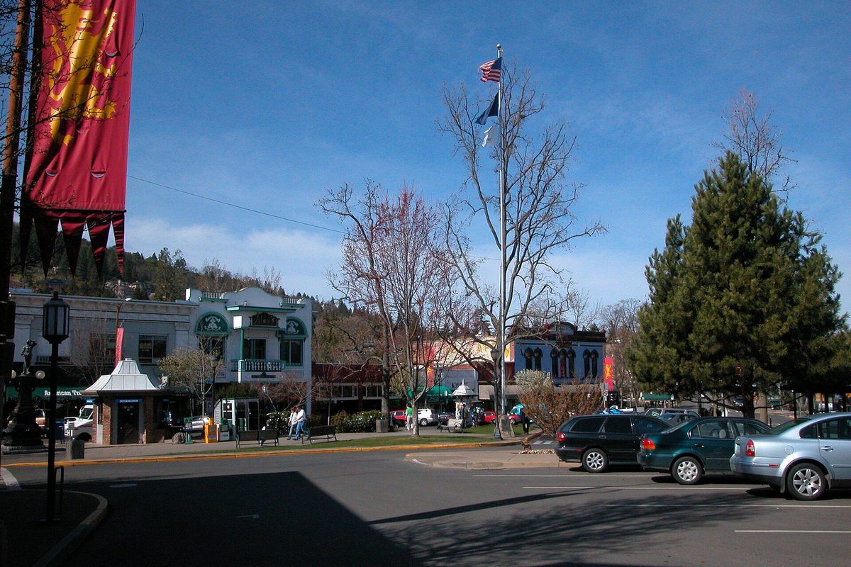 Ashland's Plaza on a clear day: shops along the street, a flagpole with the US flag, bare trees, parked cars and a red banner at the left