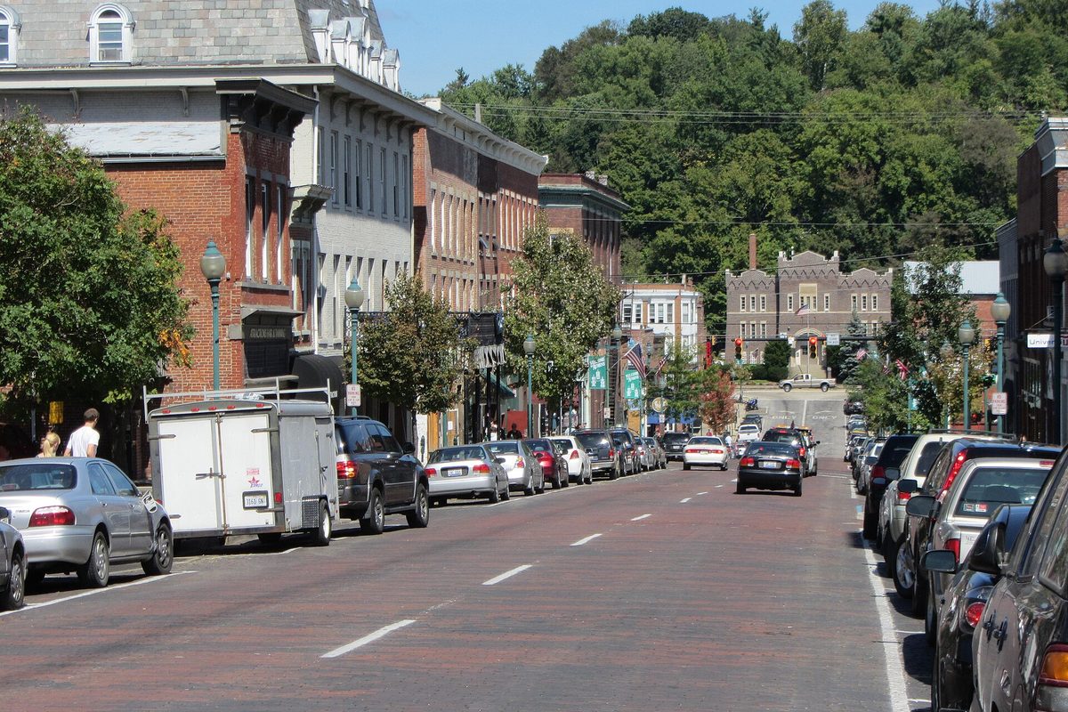A brick-paved downtown street in Athens lined with parked cars and red brick storefronts, with a wooded hillside rising behind it