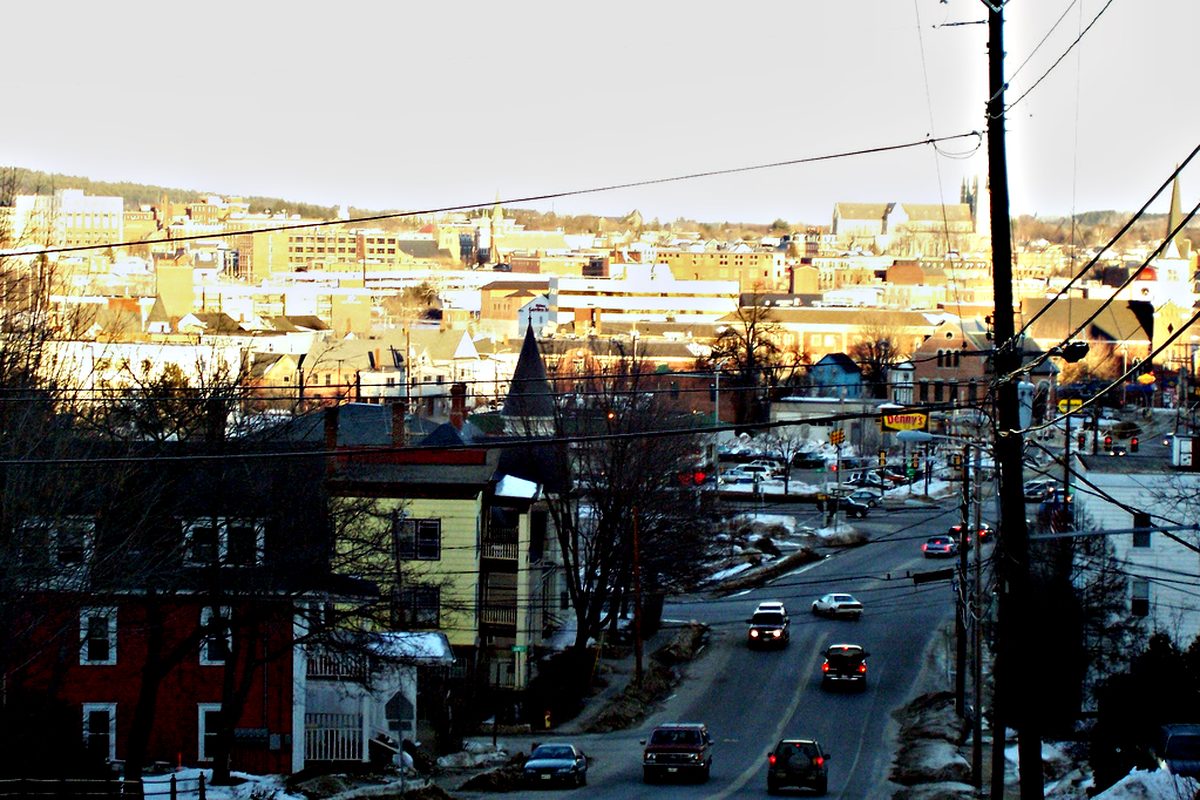 Looking down a snowy Court Street hill on a winter afternoon toward the brick buildings and steeples of Auburn and Lewiston, with cars on the road and power lines overhead