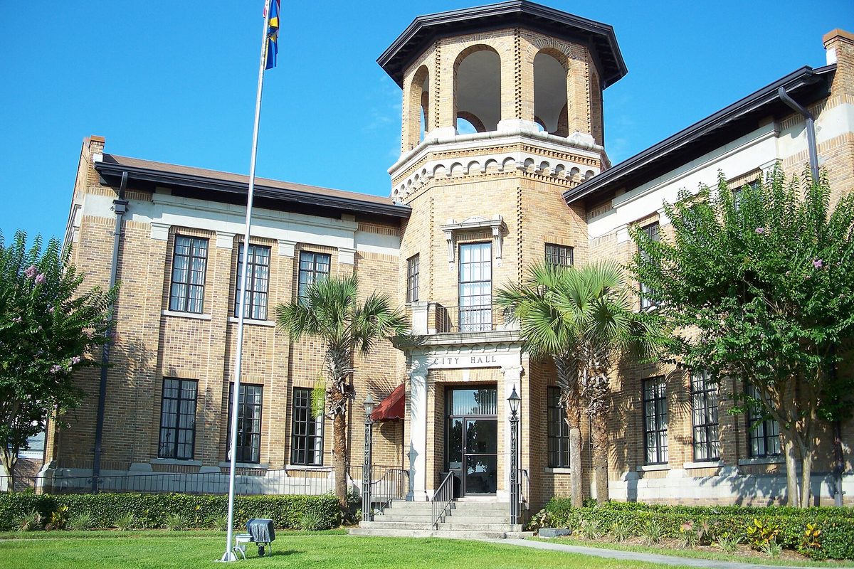 Auburndale City Hall, a two-story tan brick building with an open octagonal tower over the entrance, palm trees, a flagpole and a green lawn