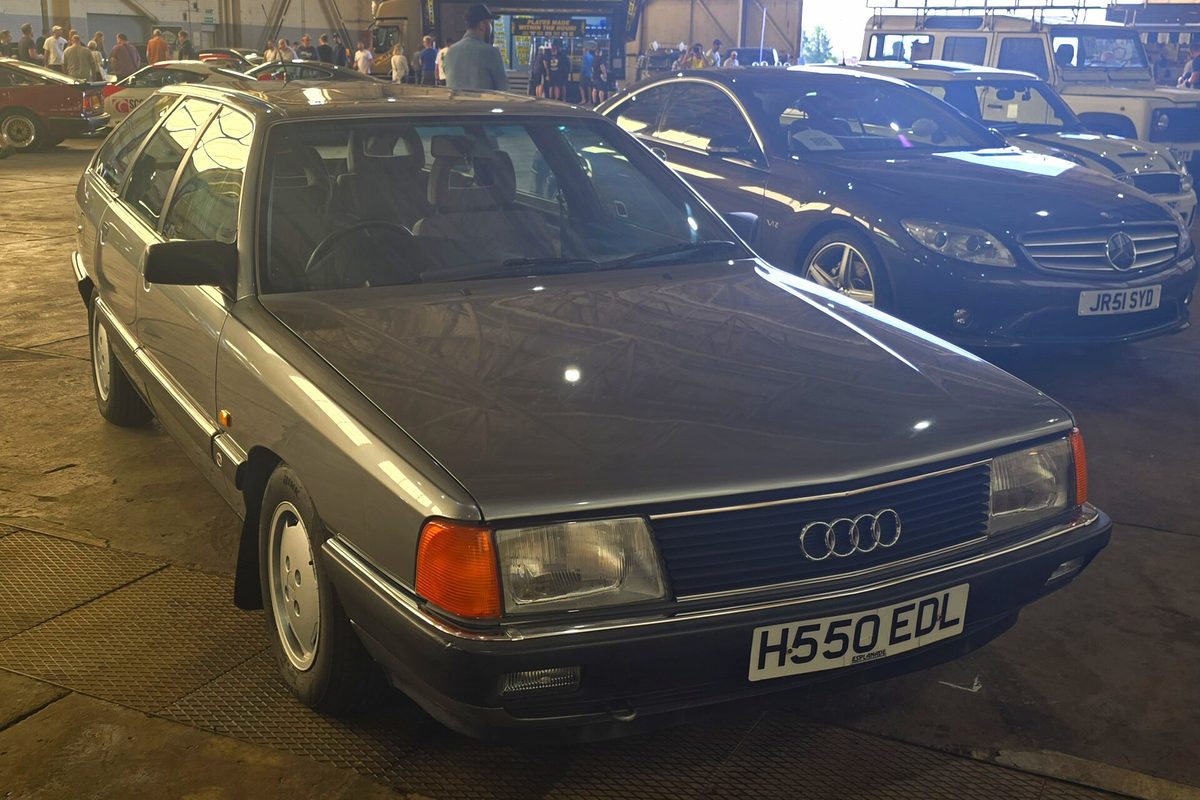A gray 1991 Audi 100 Avant wagon parked indoors at a car show, next to a black sedan