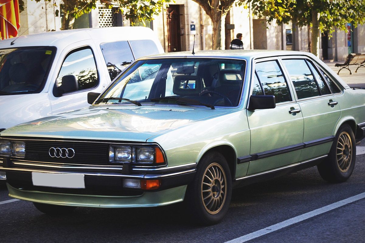 A pale green early-1980s Audi 200 sedan with gold-colored wheels, stopped on a city street beside a white van