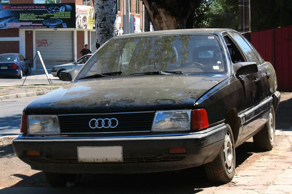A dusty black 1986 Audi 5000 S sedan parked at a curb on a sunny street, seen from the front
