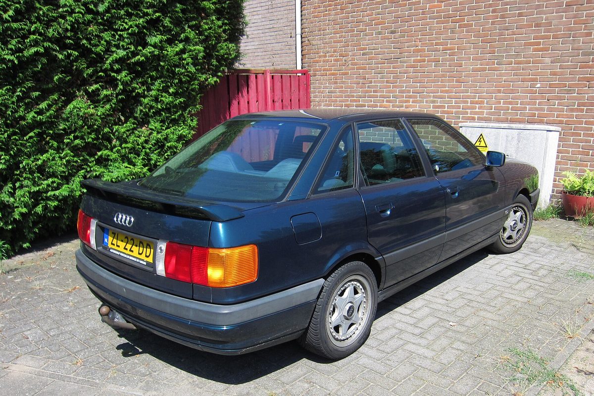 A dark blue-green Audi 80 sedan seen from the rear, parked on brick pavers beside a tall hedge