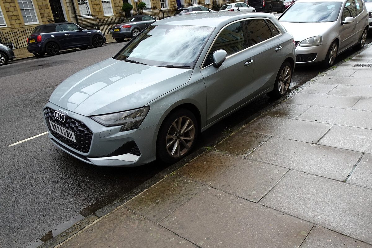 A grey-green Audi A3 Sportback hatchback parked along a wet stone curb on a city street