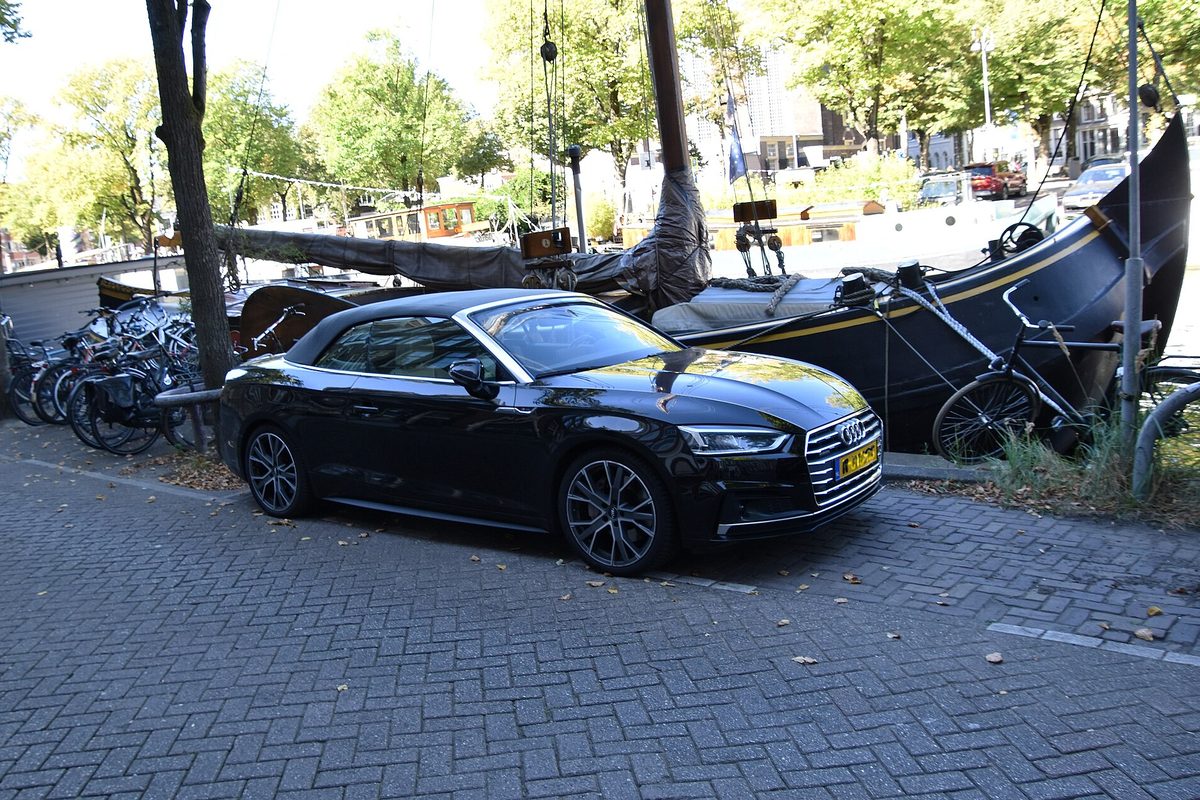 A black Audi A5 Cabriolet parked on brick pavers beside a row of bicycles and a moored canal boat