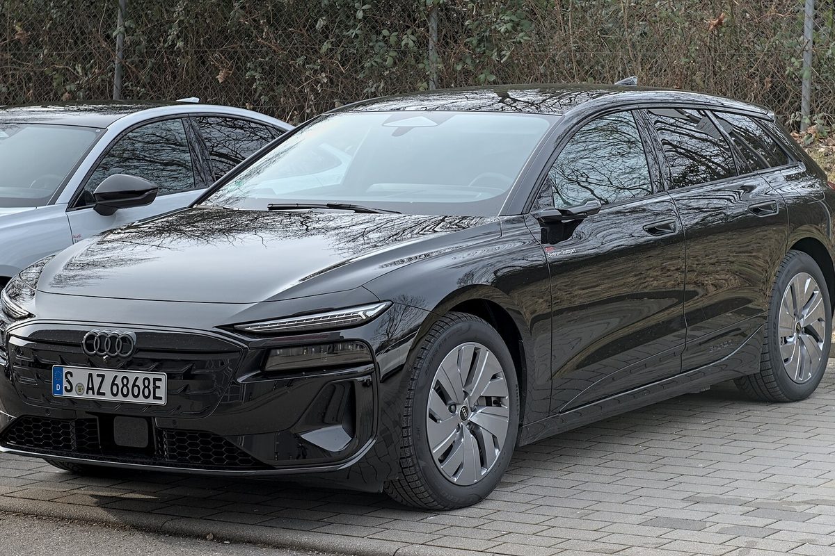 A black Audi A6 Avant e-tron wagon parked on gray paving stones beside another car, with bare hedges behind
