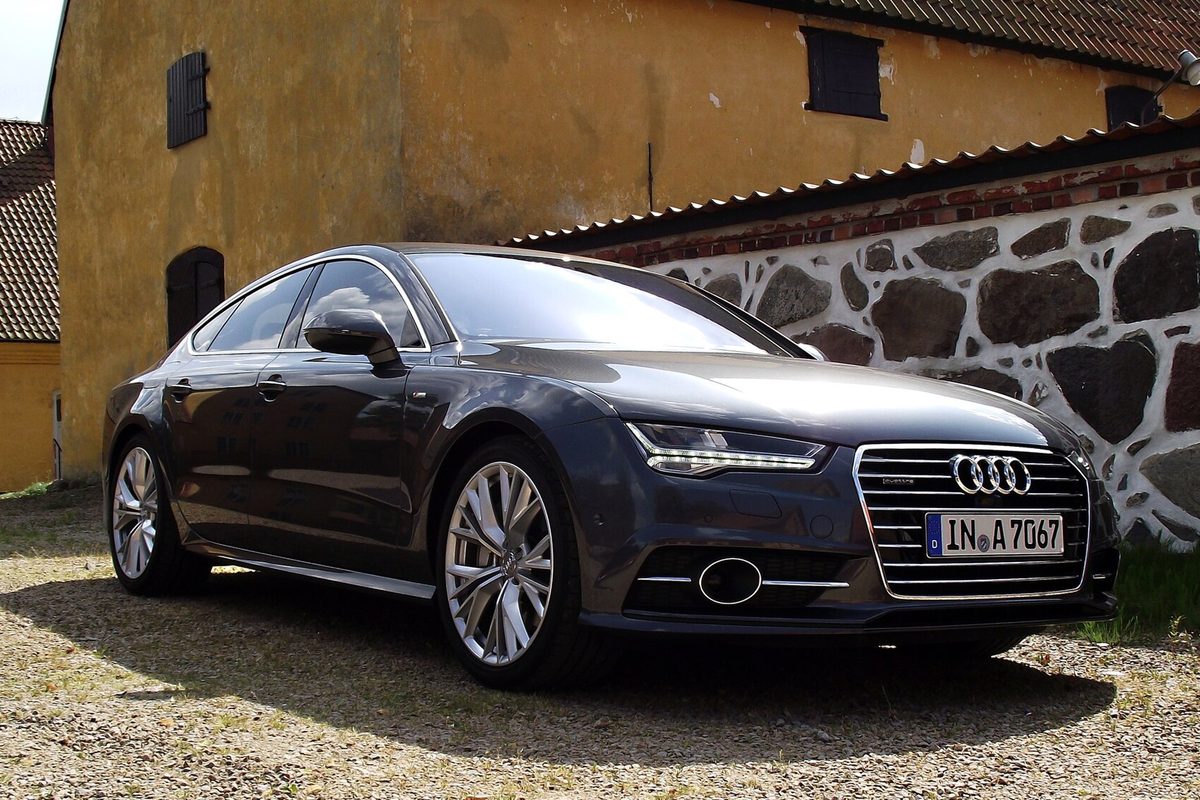A dark grey Audi A7 Sportback parked on gravel in front of a yellow farmhouse and a stone wall