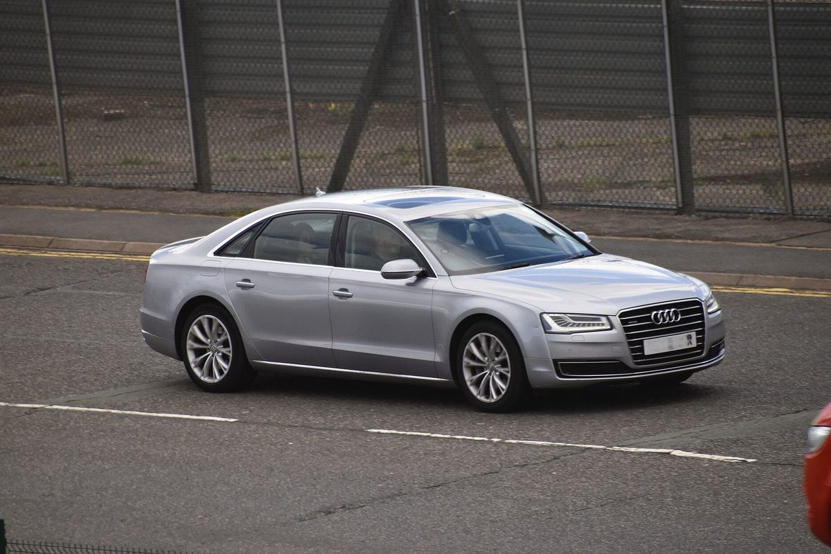 A silver Audi A8 sedan driving on a road beside a tall chain-link fence