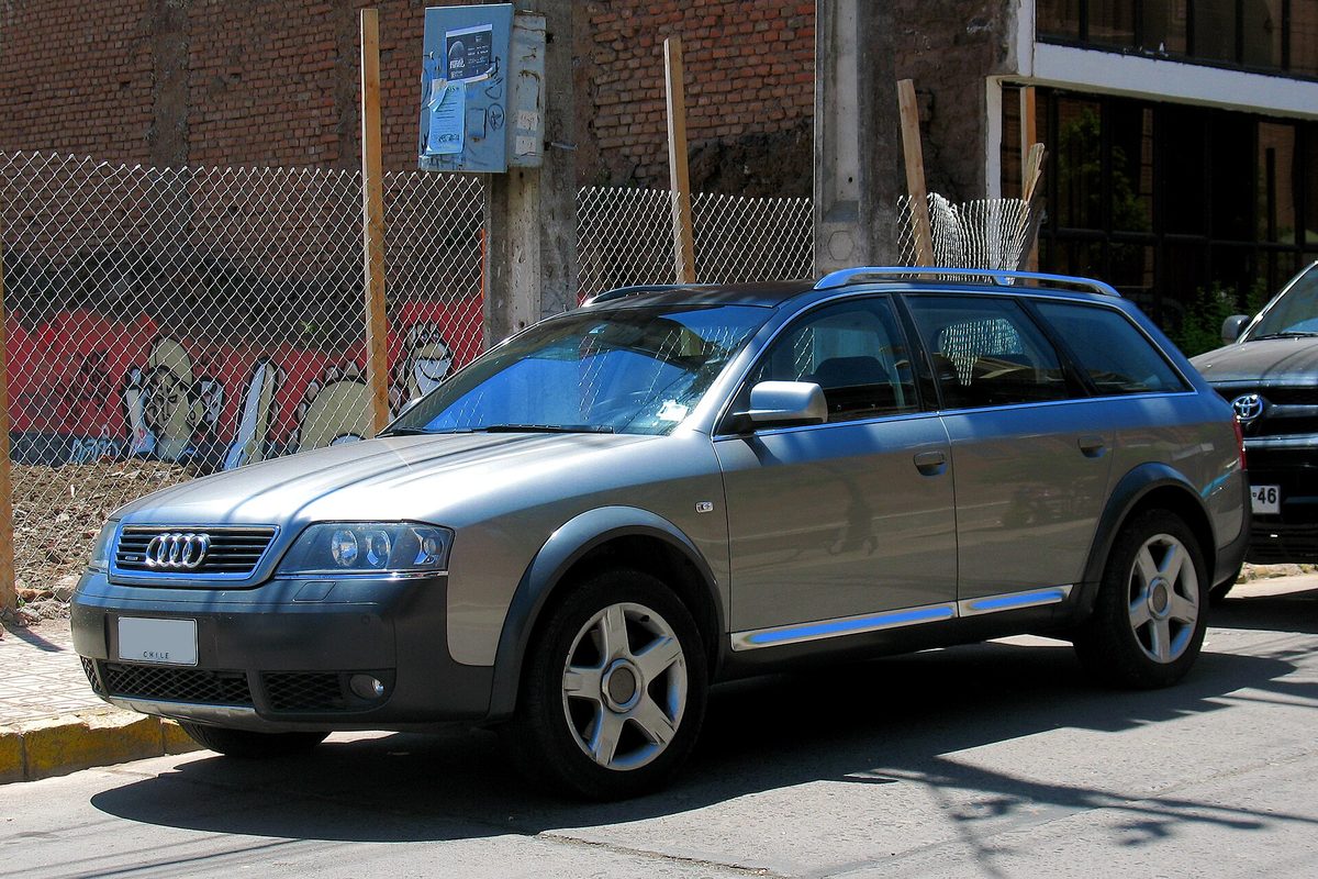 A silver two-tone 2004 Audi A6 allroad wagon with roof rails, parked on a street beside a fence and a graffiti-covered wall