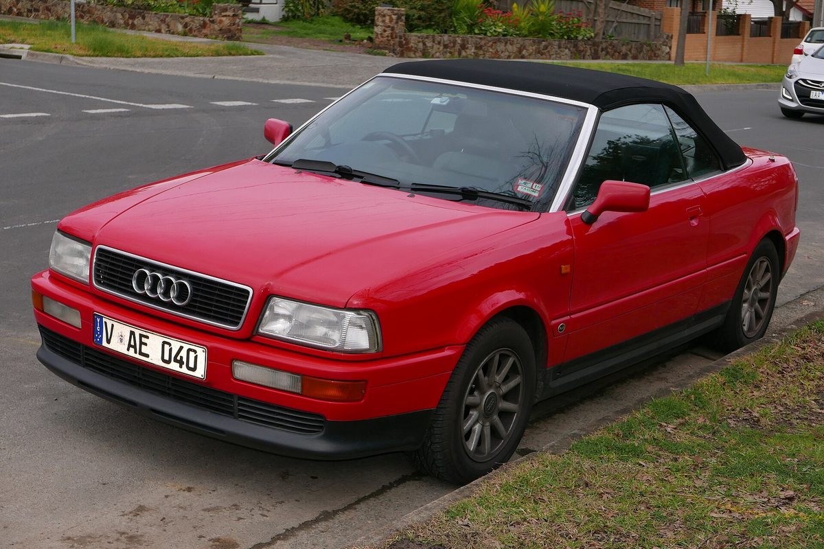 A red Audi Cabriolet convertible with its black soft top up, parked at a curb next to a grass strip