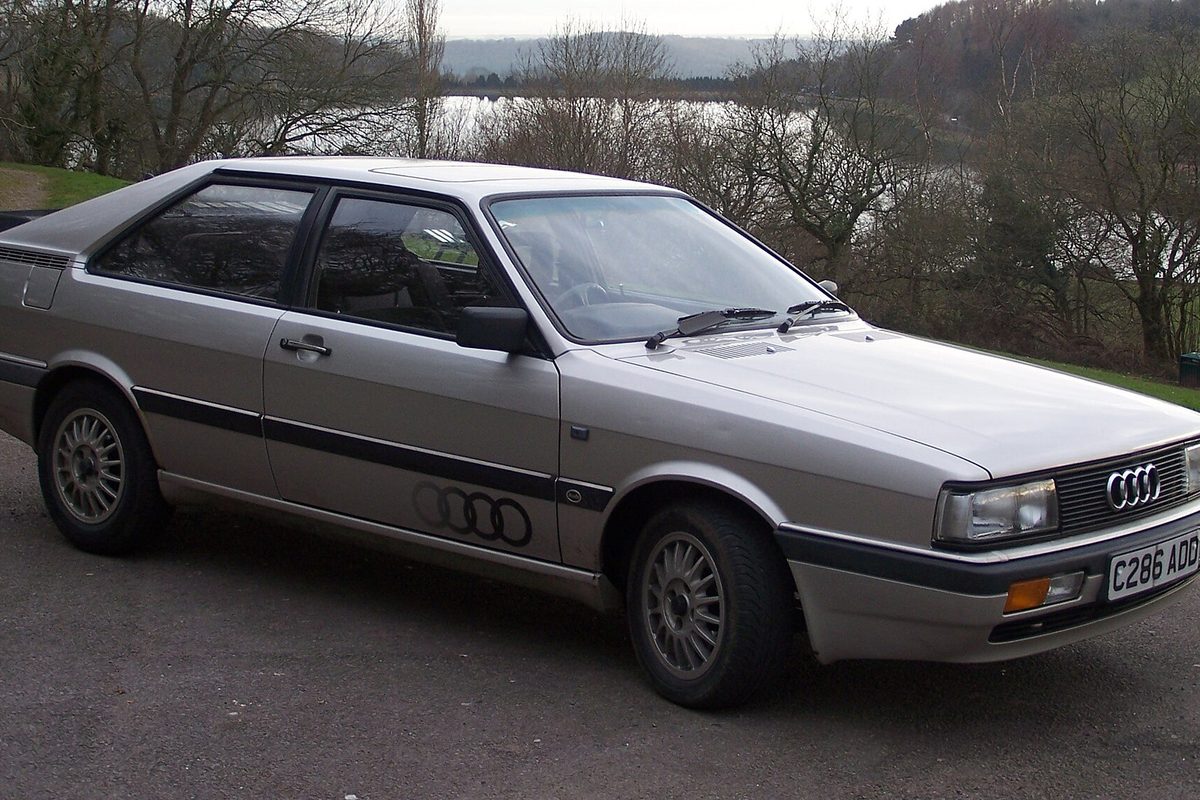 A silver-beige mid-1980s Audi Coupe GT hatchback parked on a hillside road with bare trees and a lake behind it