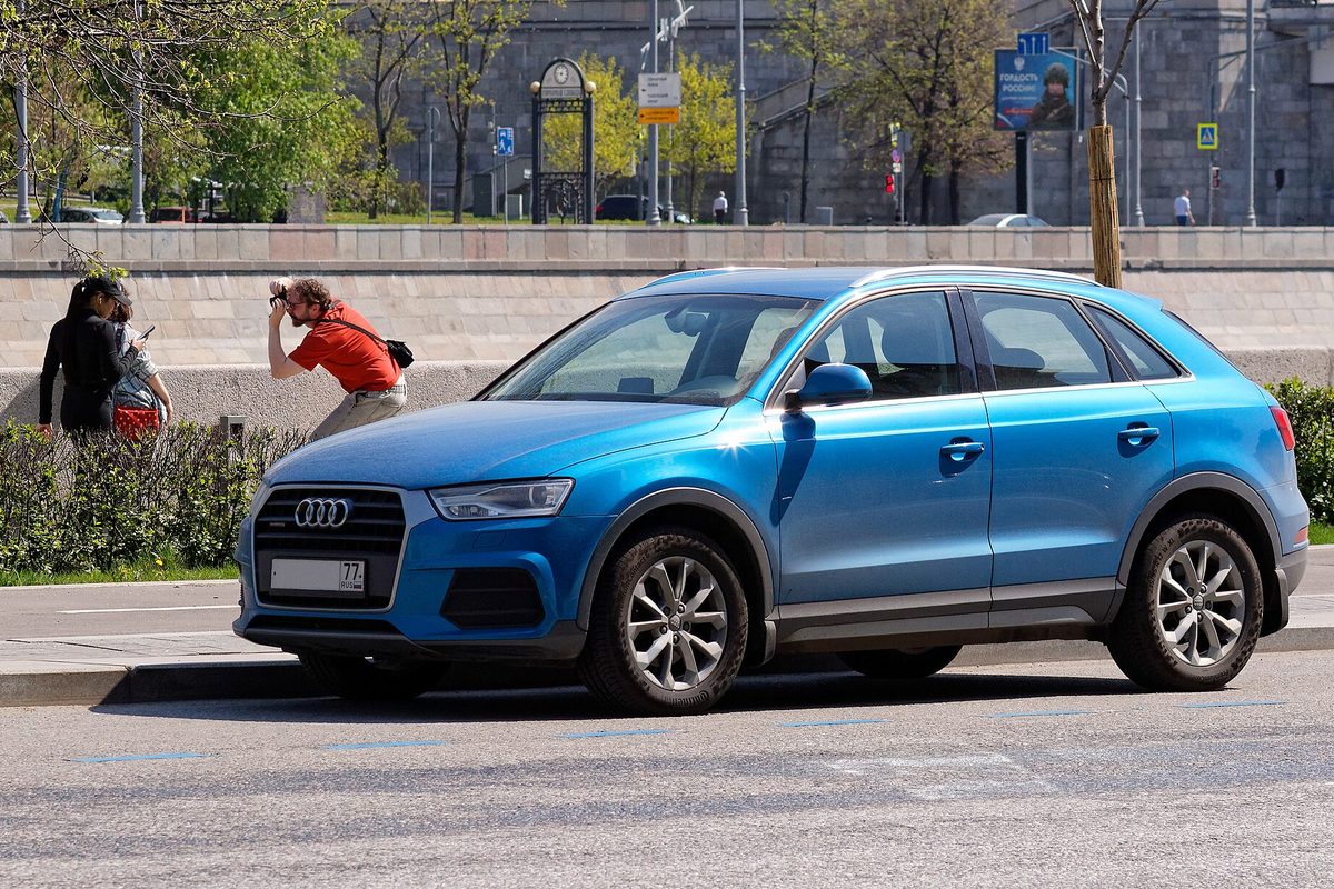 A bright blue Audi Q3 parked at a city curb on a sunny day, with people on the sidewalk behind it