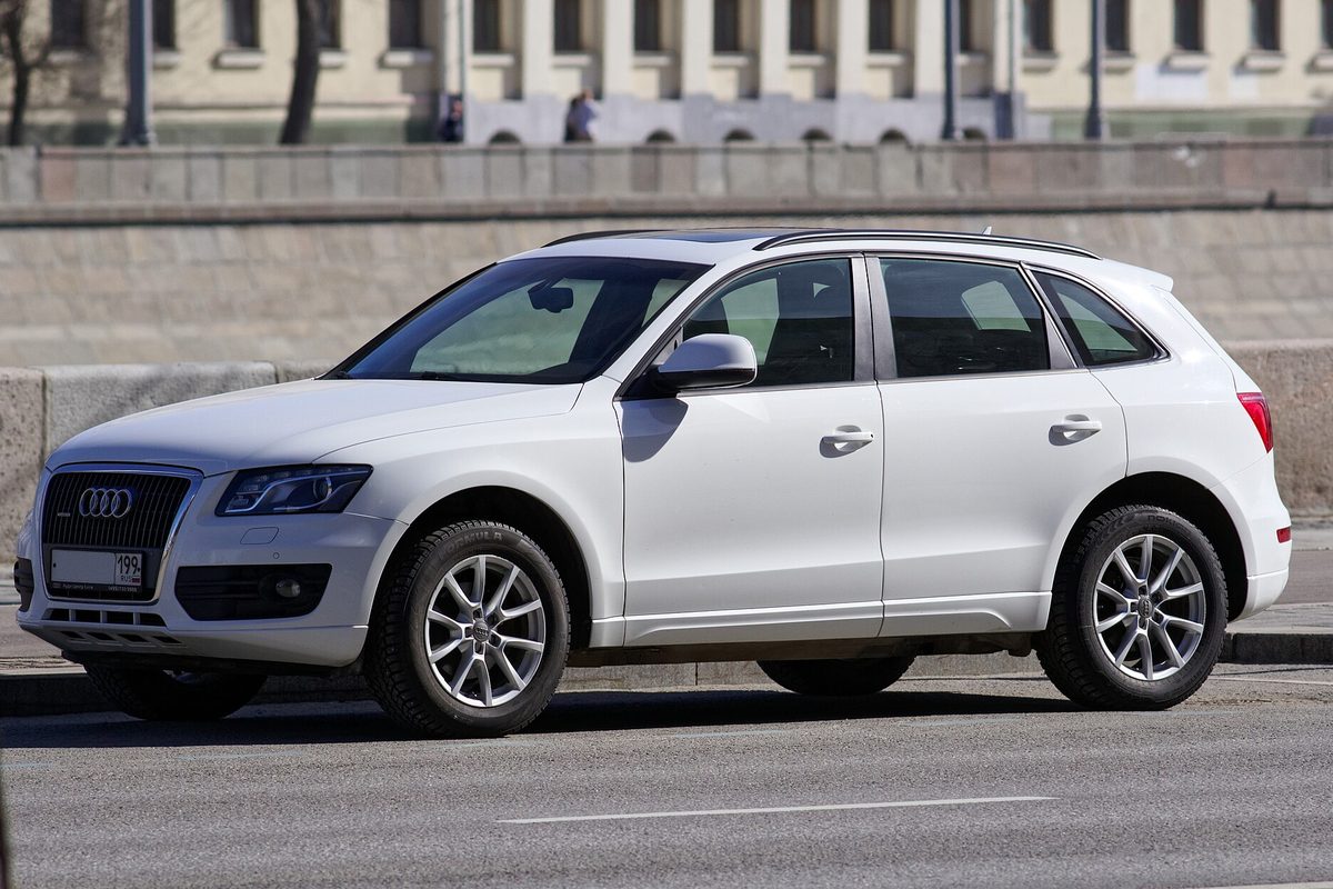 A white first-generation Audi Q5 parked on a city street in front of a stone embankment wall