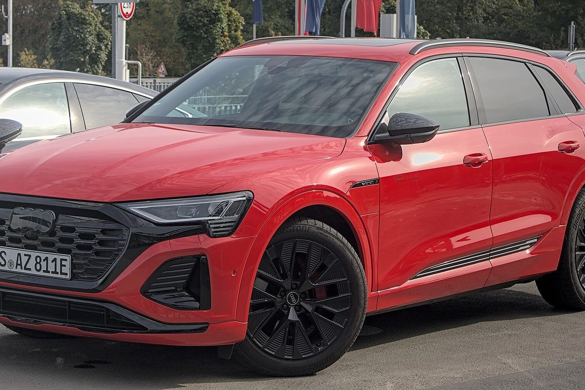 A red Audi Q8 e-tron SUV with black wheels and black trim, parked in a lot with flags and trees behind