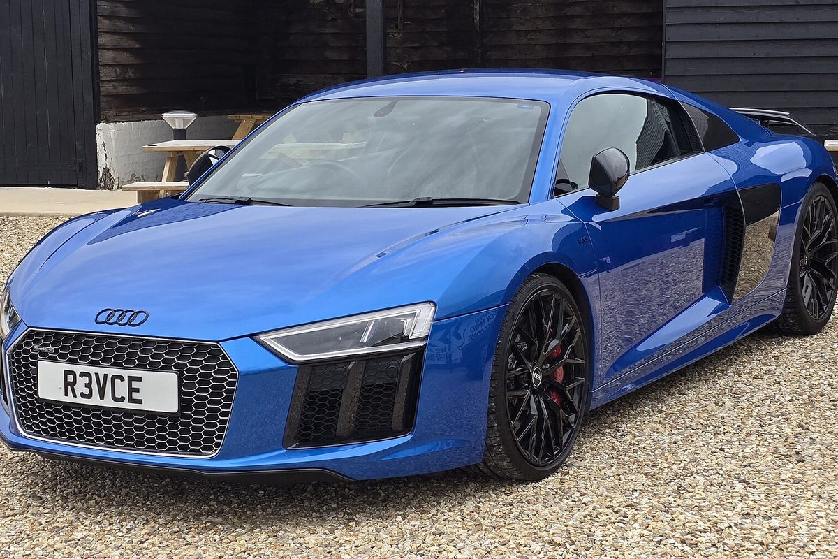 A bright blue Audi R8 coupe with black wheels, parked on gravel by a dark wooden building