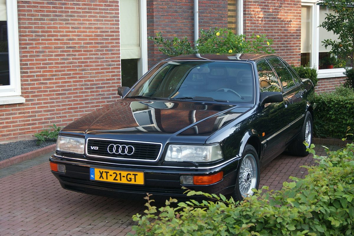 A black Audi V8 sedan with a chrome V8 grille badge and wire-spoke wheels, parked on a brick driveway in front of a brick house