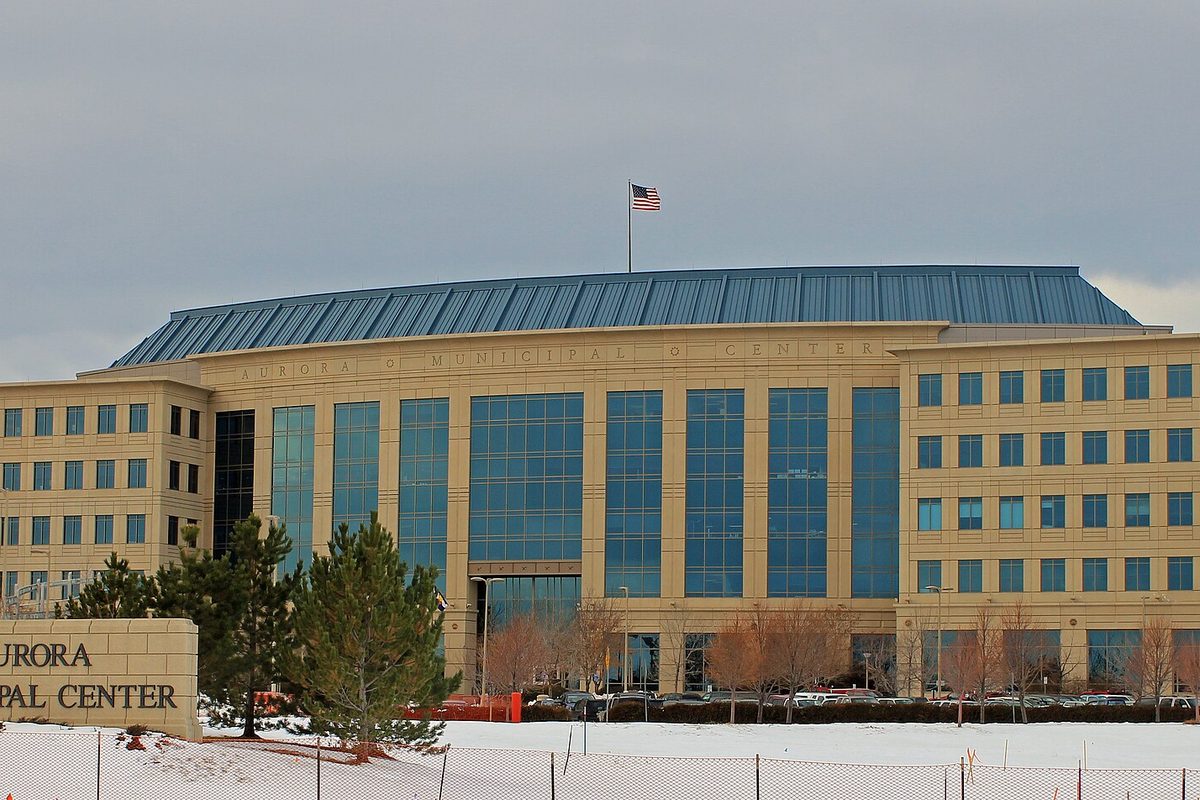 The Aurora Municipal Center, a tan stone and blue glass building with a curved roof and a flag, behind snow-covered ground