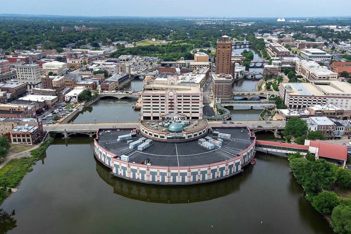 Aerial view of downtown Aurora on the Fox River, with a large round building on the water, several bridges and a tall brick tower