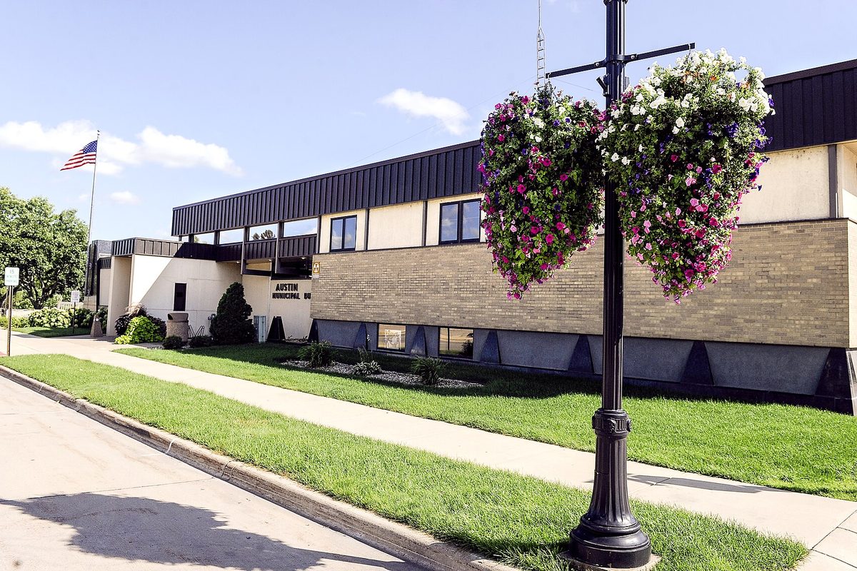 The Austin Municipal Building, a low tan-brick city building with a dark upper band, with baskets of pink and white flowers hanging from a lamp post and a flag out front
