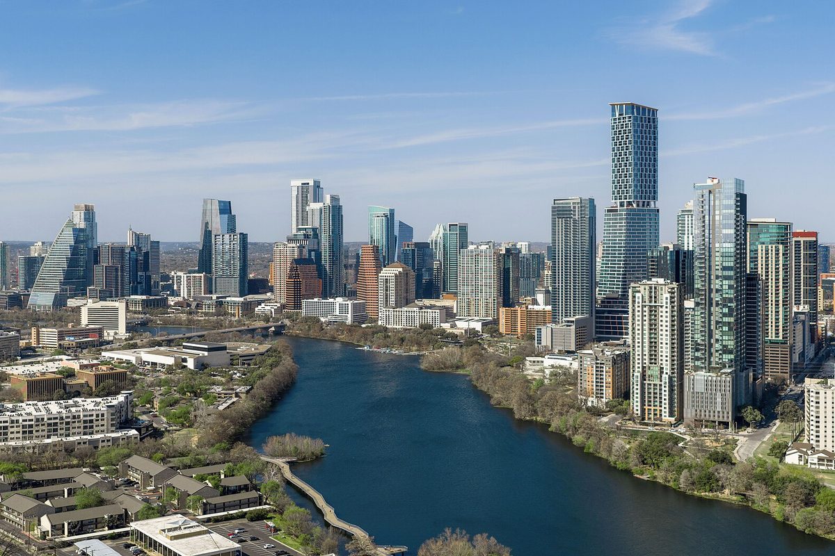 Downtown Austin's cluster of glass high-rises on a clear day, seen from above a wide river lined with trees, with low-rise buildings in the foreground