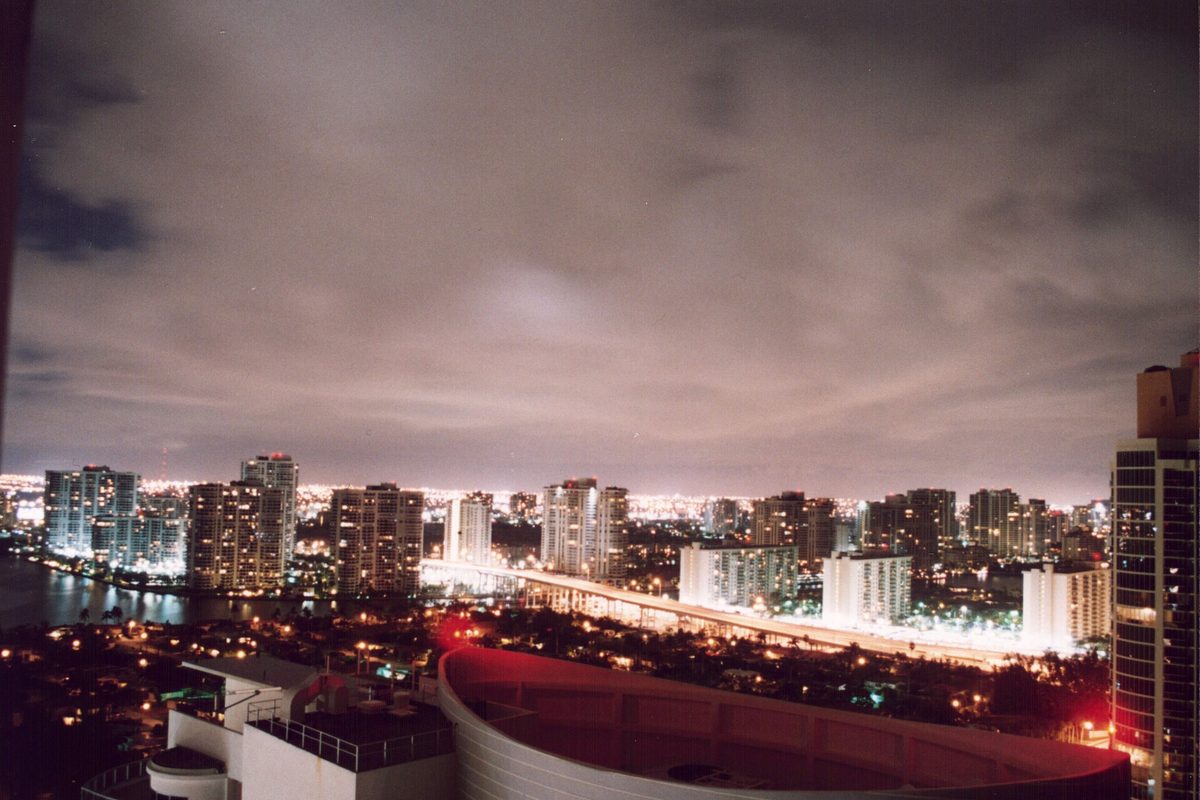Aventura at night seen from a high floor: lit residential towers, a bright road running along the water and a low cloudy sky