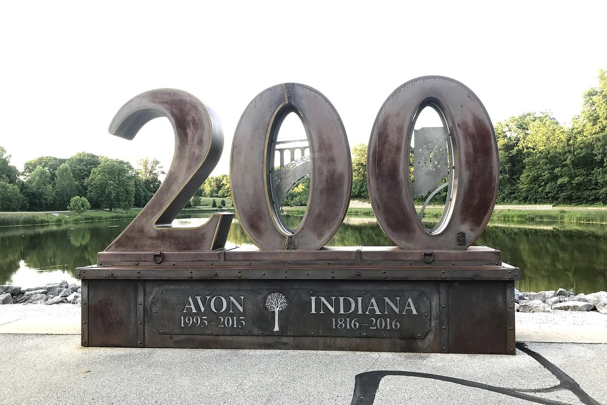 A large rusted-metal sculpture of the number 200 with Avon and Indiana plaques on its base, standing in front of a pond and green trees