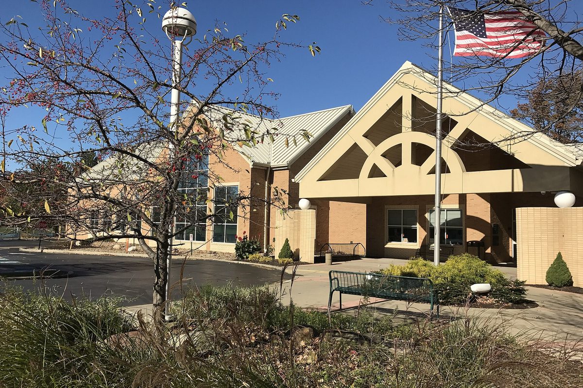 The Avon Lake Public Library, a tan-brick building with a gabled entrance canopy, a flagpole flying the American flag, a green bench and ornamental grasses under a blue sky