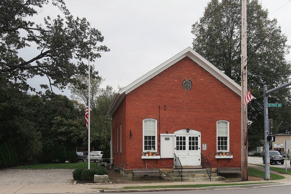 Avon's original town hall, a small red-brick building with white double doors and American flags, on a corner beside a Detroit Rd street sign