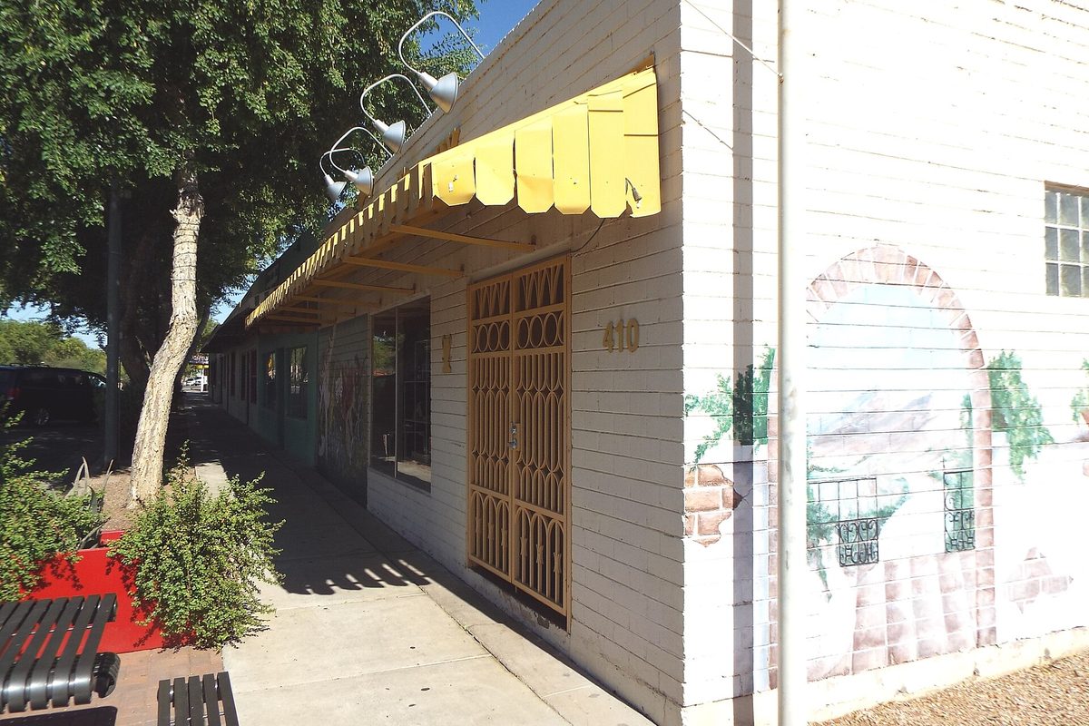 A white-painted building in historic downtown Avondale with a yellow scalloped awning, an ornate gold door grille and a painted arch mural, beside a shaded sidewalk
