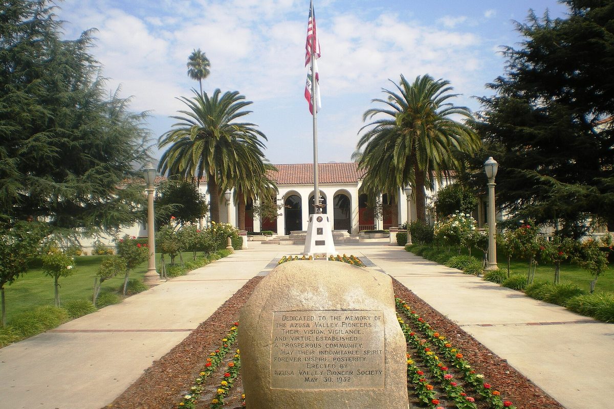 Azusa City Hall, a Spanish-style building with a red tile roof at the end of a walkway lined with palms, with a flagpole and a carved stone memorial to Azusa Valley pioneers in a flower bed