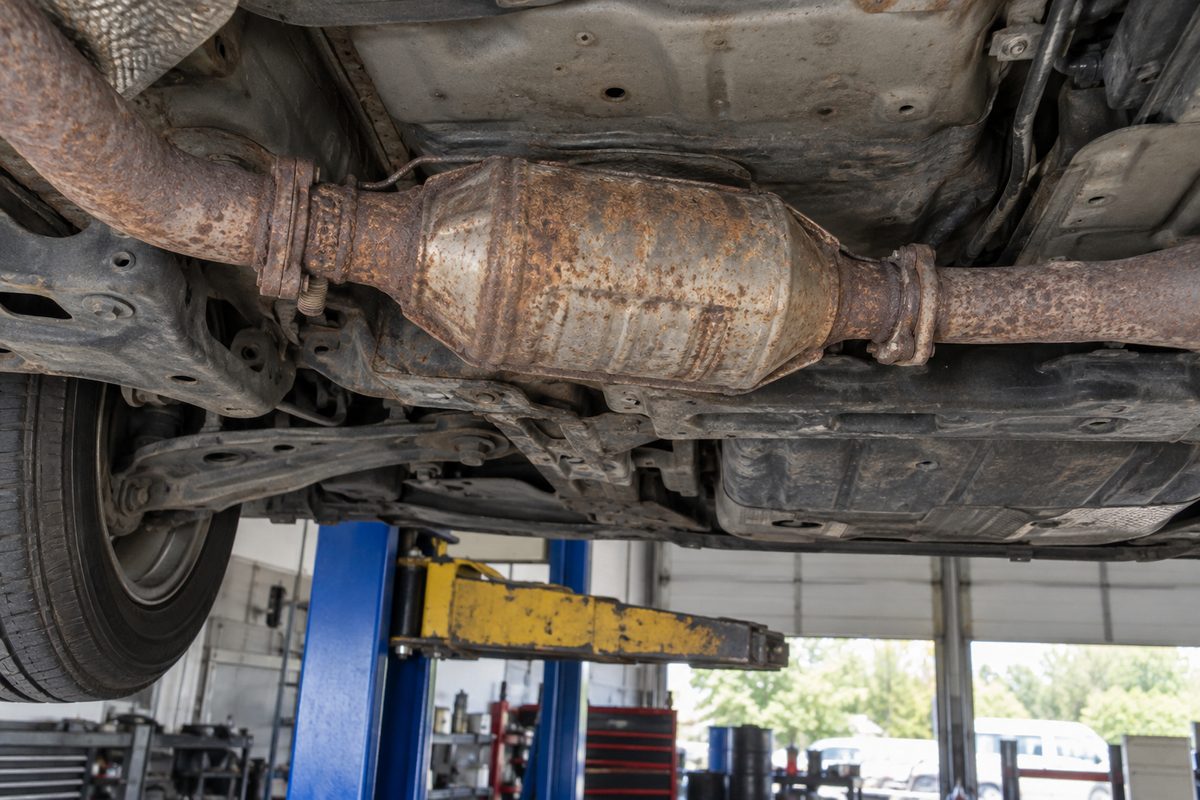 A rusty catalytic converter and exhaust pipe seen from below a car raised on a blue two-post lift in a repair shop