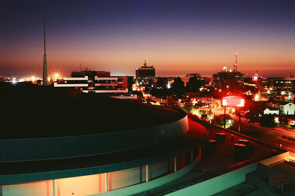 Downtown Bakersfield at dusk, office towers and a radio mast against an orange sky, with the curved roof of the arena in the foreground