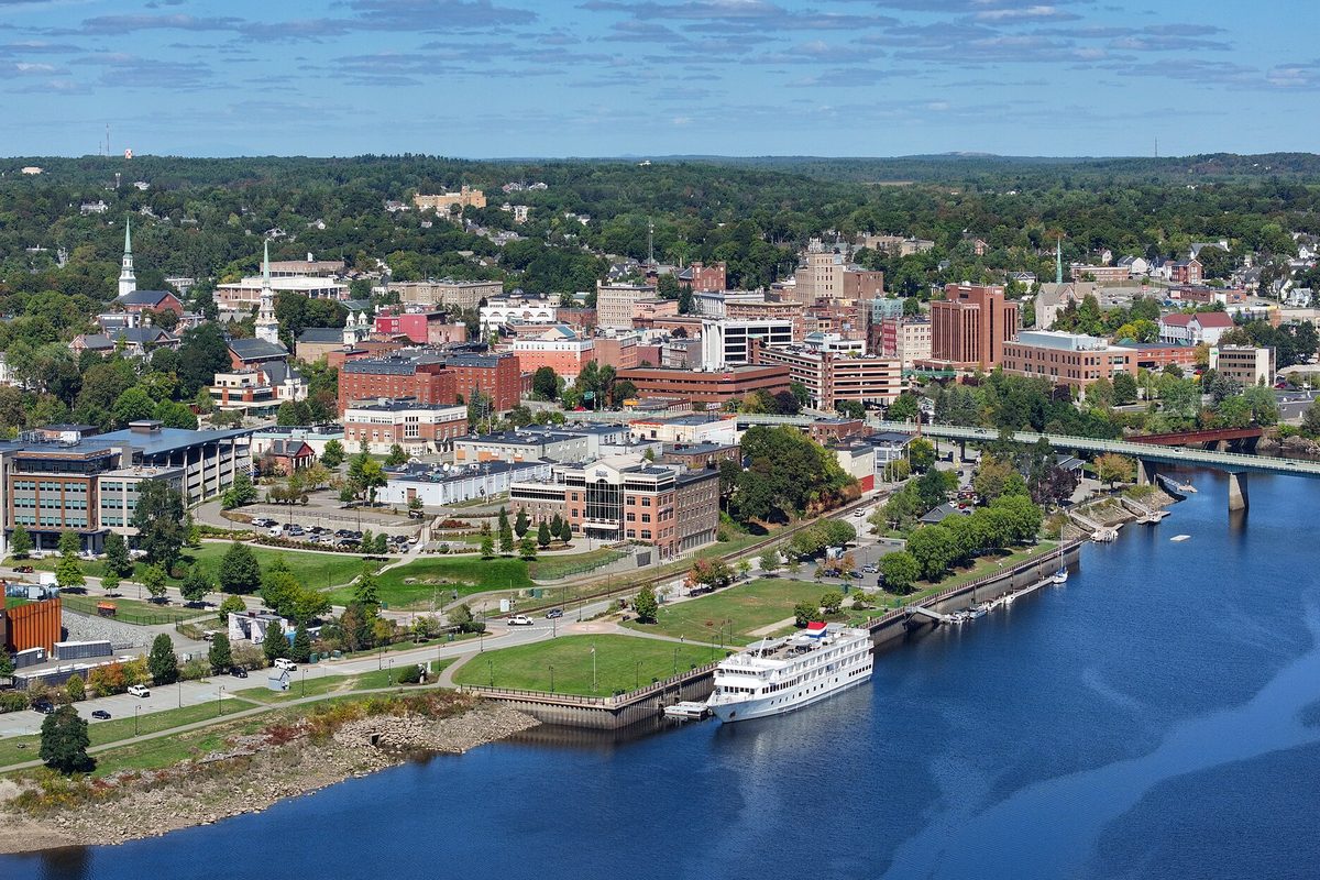 Aerial view of downtown Bangor beside a wide river, with a white riverboat docked at a waterfront park and a bridge crossing the water on the right