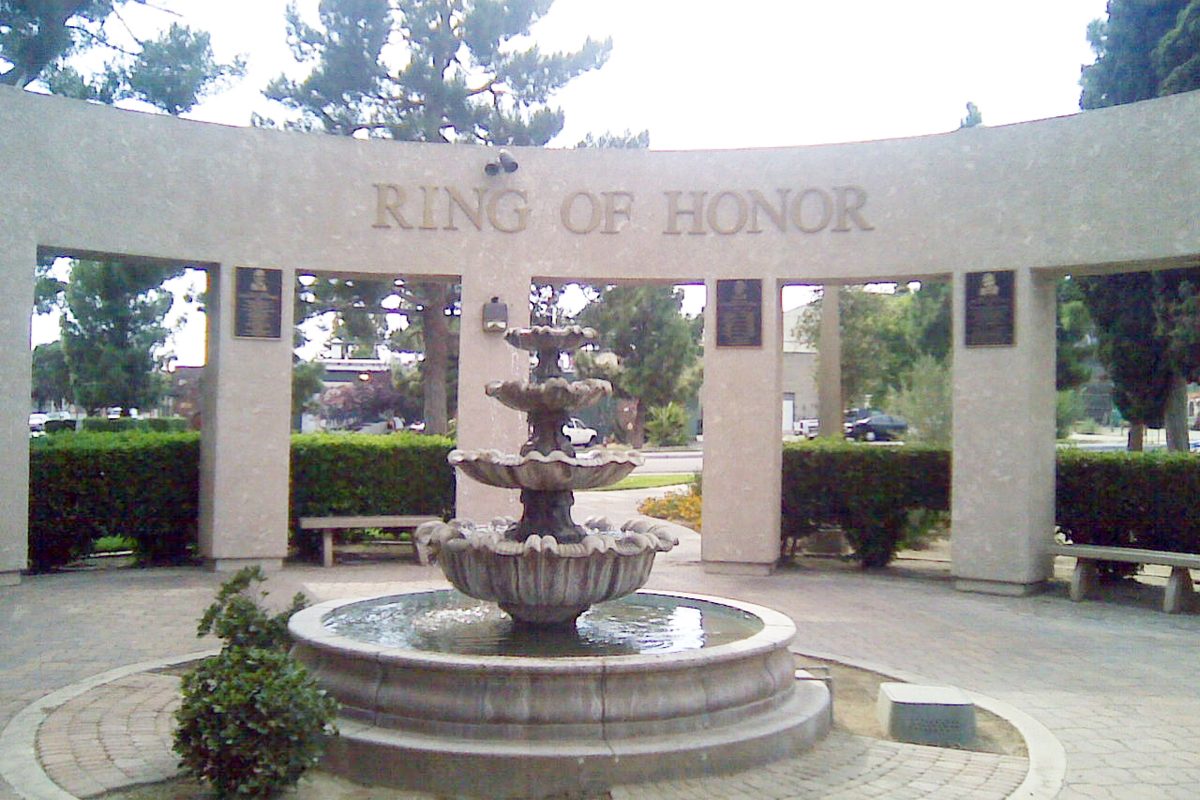A tiered stone fountain in front of a curved arch lettered Ring of Honor, with plaques on its pillars, hedges and pine trees in Banning