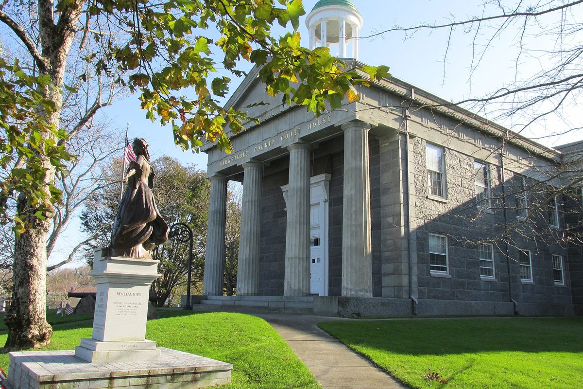 The gray granite Barnstable County Courthouse with columns and a white cupola, a bronze statue on a pedestal in front and autumn leaves overhead