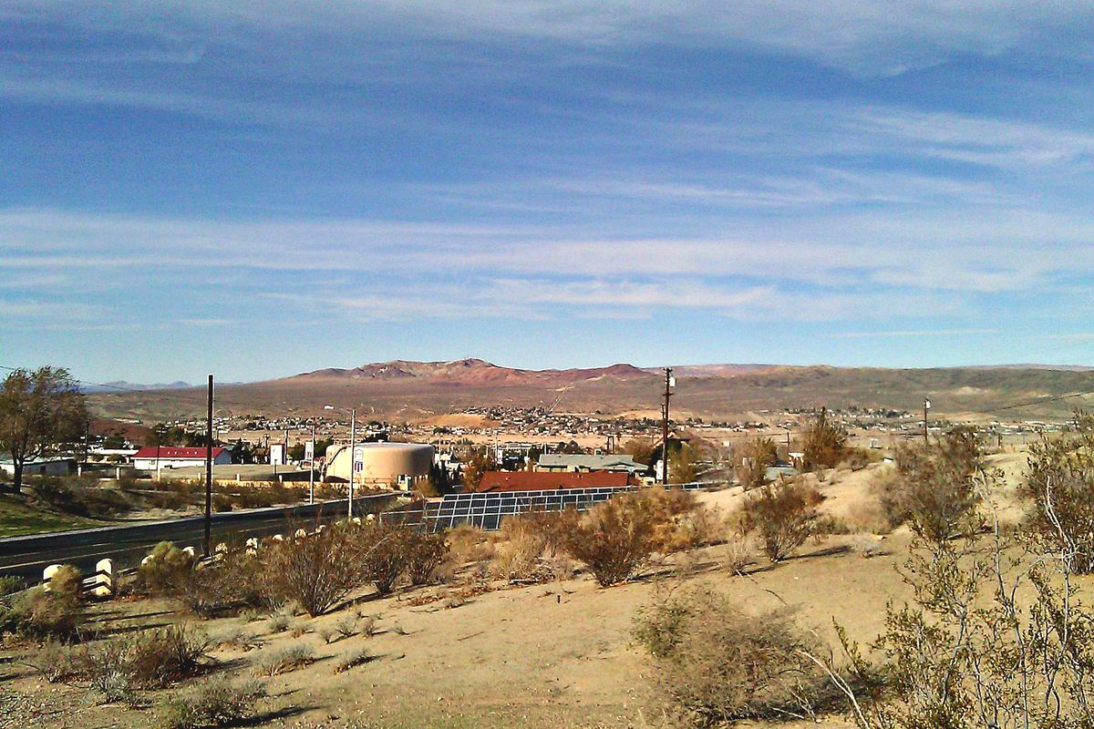 Desert brush on a slope above Barstow, with a round water tank, rooftops and red-brown mountains under a streaked blue sky