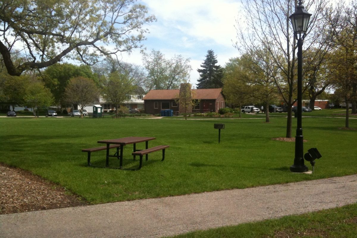 A green lawn in Bartlett Park with a picnic table, a black lamp post and a path, a small brick building behind spring trees