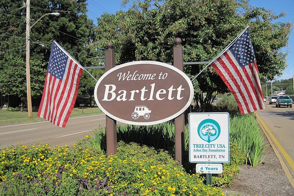 A brown oval Welcome to Bartlett sign between two American flags, with yellow flowers in front and a green Tree City USA sign beside it