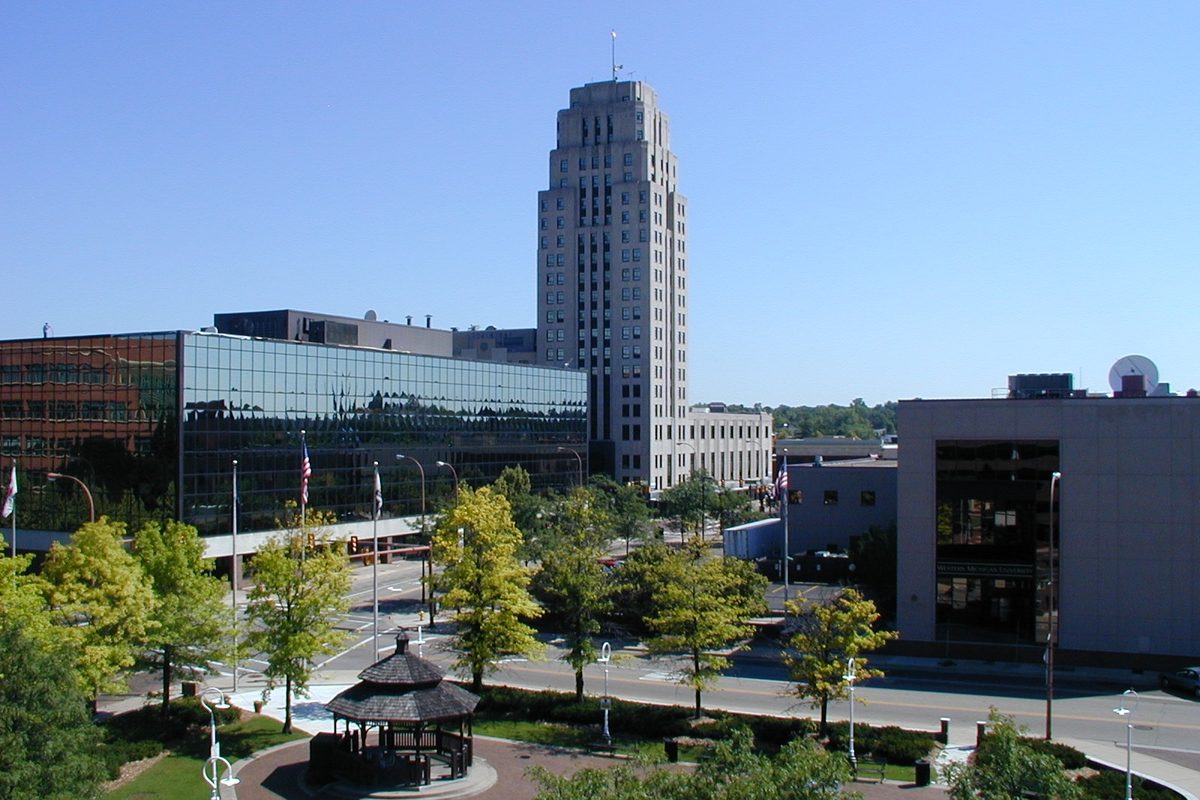 Downtown Battle Creek under a clear blue sky, with a stepped stone tower, a glass office building and a gazebo in a small tree-lined park