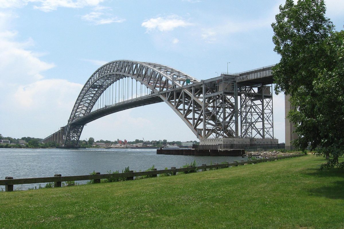 The steel arch of the Bayonne Bridge over the water, seen from the lawn and wooden fence of Collins Park in Bayonne