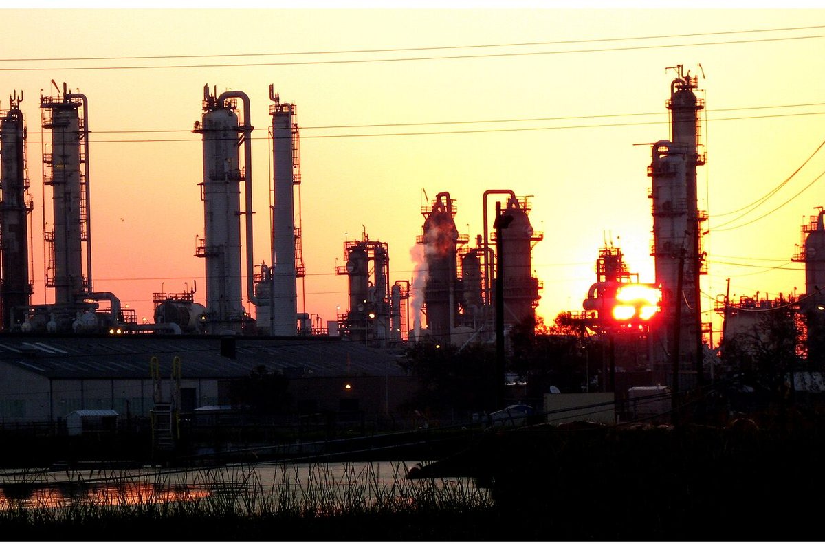 Towers and stacks of a chemical plant near Baytown in silhouette against an orange sunrise, with marsh grass and still water in front
