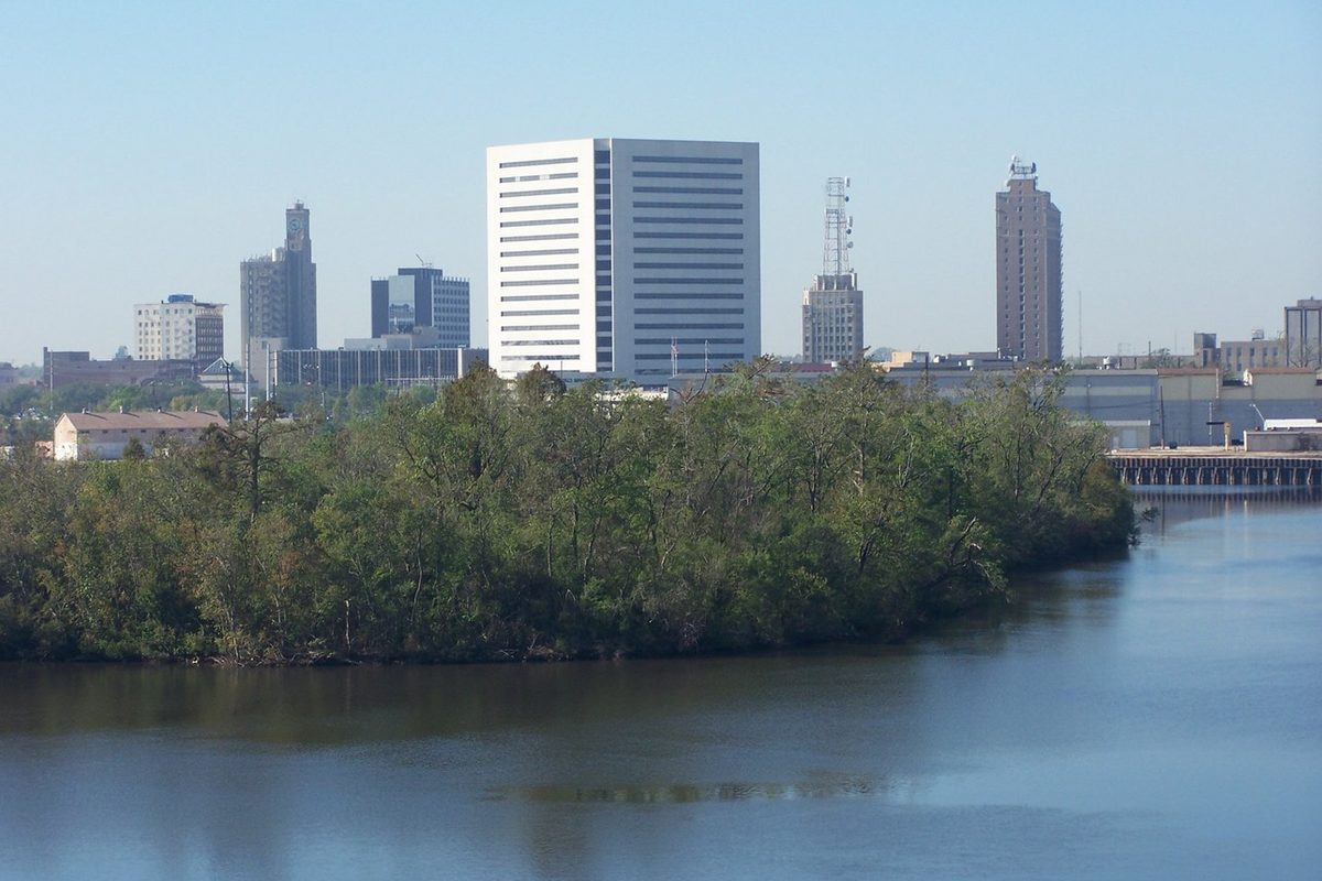 Downtown Beaumont's office towers, led by a tall white high-rise, rising behind a thick stand of trees on a riverbank, seen across calm blue water