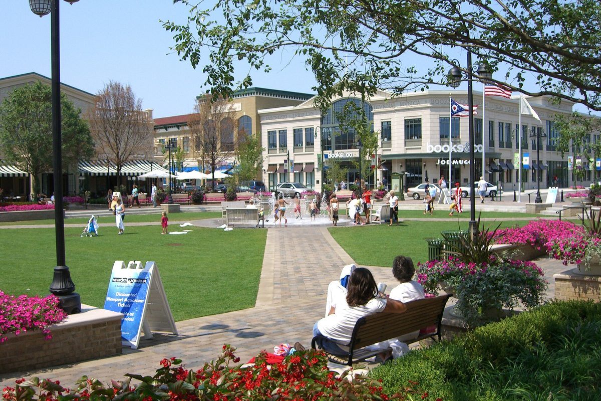 Families at the splash fountain on the lawn at The Greene Town Center in Beavercreek, with flower beds, a bench and storefronts behind