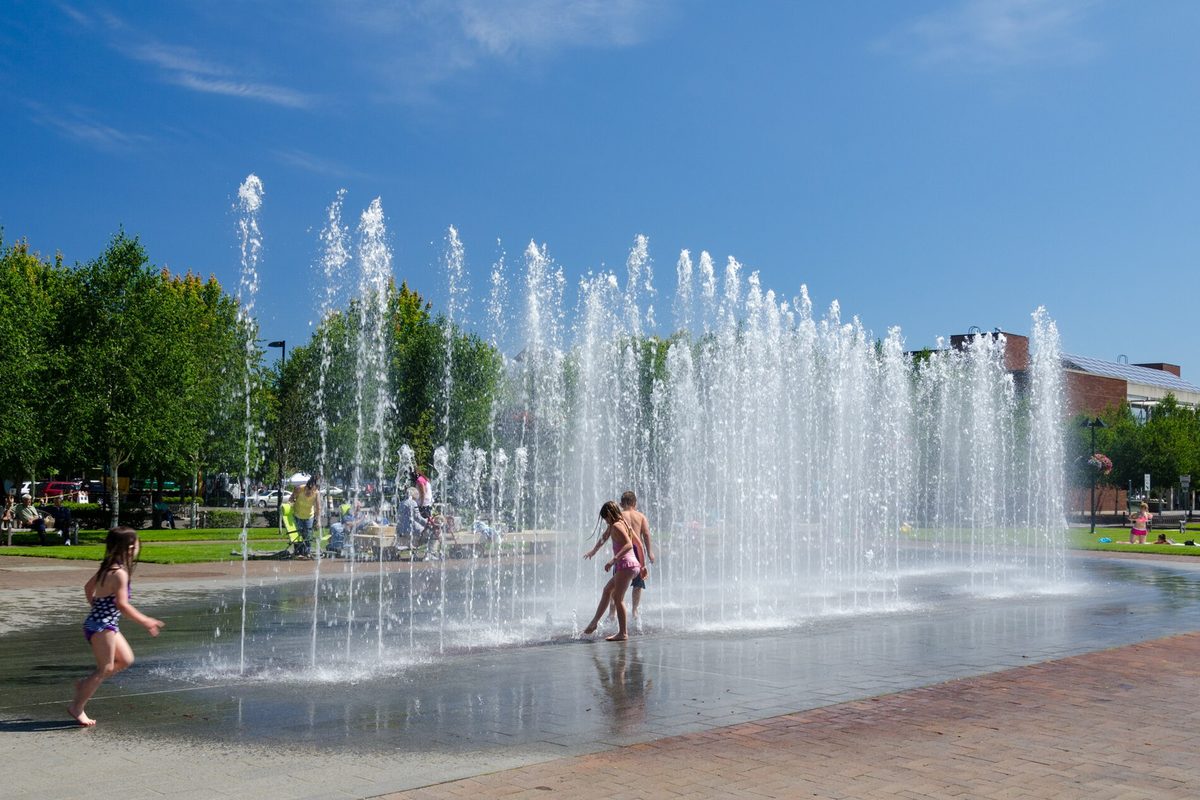 Children playing in the jets of the fountain at Beaverton City Park on a sunny day, with trees and a brick building behind