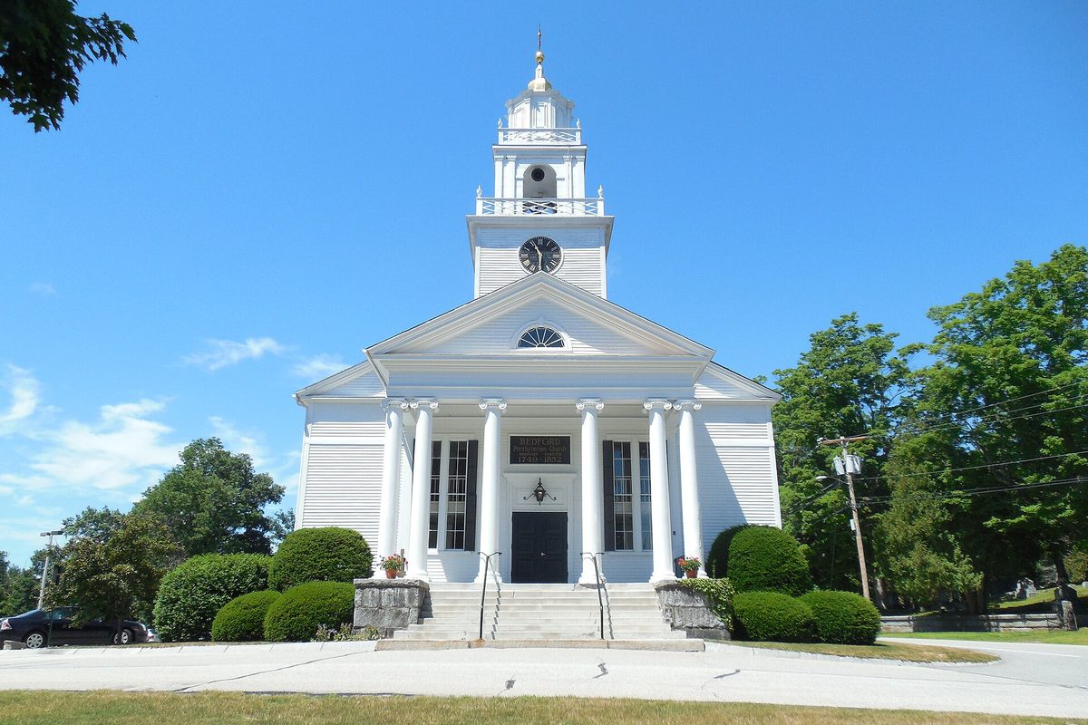 The white Presbyterian Church in Bedford, with tall columns, a clock tower topped by a steeple, and trimmed shrubs along the front