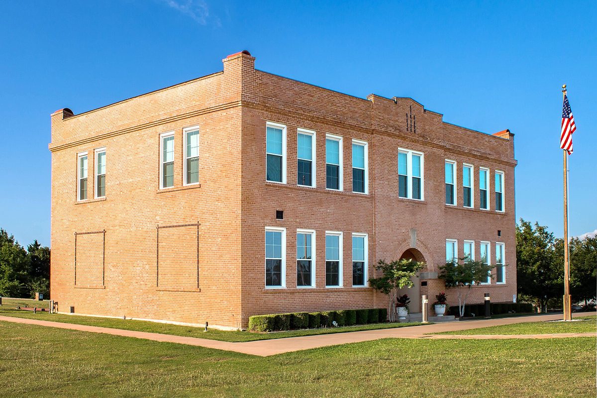 The two-story red brick Bedford School building on a green lawn under a clear blue sky, with an American flag on a tall pole